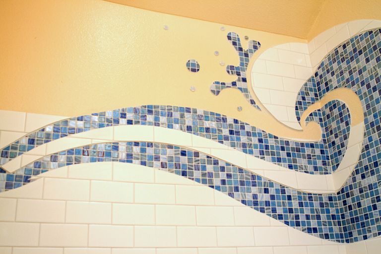 Shower wall with a Hokusai-style cresting blue glass mosaic ocean wave and droplet spray pattern, set against white subway tile and pale yellow paint
