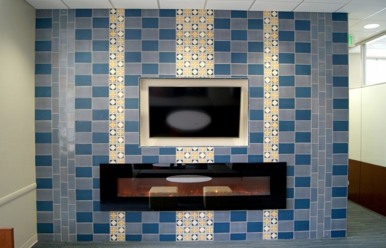 Commercial media wall clad in Rookwood tile — alternating blue and slate gray field with vertical decorative inlay columns of cream and gold quatrefoil-pattern tiles, surrounding a mounted TV and linear gas fireplace